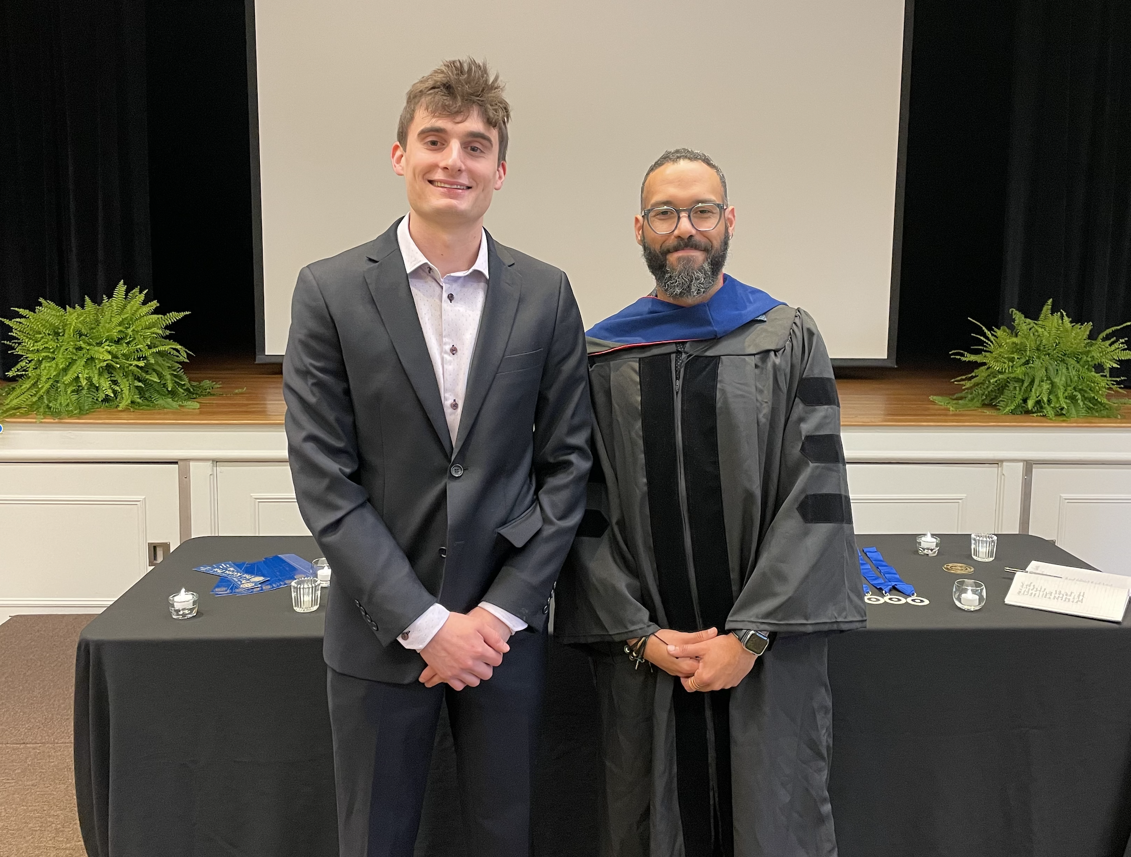 A student in a suit and a faculty member in academic regalia smile together in front of a presentation screen at an awards ceremony.